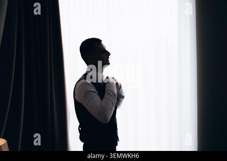 Silhouette eines Mannes, der seine Krawatte anpasst. Er steht in der Nähe eines Fensters mit sanftem Licht. Seine Haltung deutet auf eine ruhige Konzentration hin. Ideal für Geschäftsreisen, Hochzeiten oder Formalitäten Stockfoto