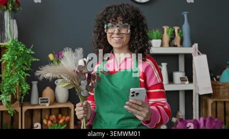 Floristin in grüner Schürze, die einen getrockneten Blumenstrauß hält und ihr Smartphone zwischen Vasen und Regalen in einem Blumengeschäft ansieht; fröhliche lokale Busi Stockfoto