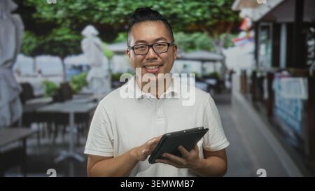 Mann, chinesisch und jung, lächelnd und klopfend mit beiden Händen auf einer Restaurantterrasse an der Straße, während er Brille und ein weißes Polo trägt; CAL Stockfoto