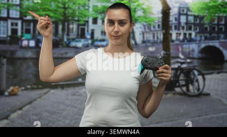 Eine Frau mit Gamepad zeigt mit dem Finger zur Seite auf einer Straße an einem Kanal mit Fahrrad und Brücke und Gebäuden, trägt weißes T-Shirt und Pferdeschwanz; Konzentration Stockfoto