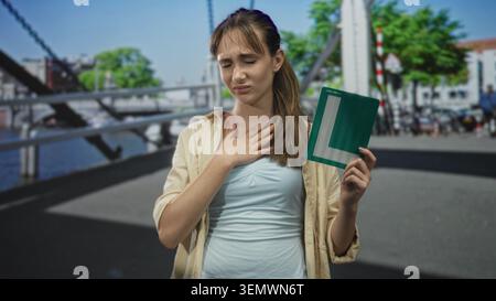 Junge Frau, die auf einer befahrenen Straßenbrücke in der Nähe des Kanals die Hand an die Kehle drückt, sichtbares Unbehagen und Lernschild; Lernstress. Stockfoto