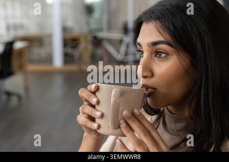 Indische asiatische Frau mit geripptem beigefarbenen Keramikbecher in der Nähe des Mundes im Büro, Kopierraum Stockfoto