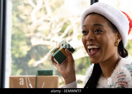 Afroamerikanerin mit grünem Geschenkkarton, Weihnachtsmann-Hut-Fuzzy-Pullover am Fenster, Kopierraum Stockfoto