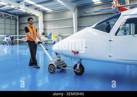 Flugzeugtechniker mit Sicherheitsweste schiebt ein Flugzeug mit einem elektrischen Schlepper, bewegt das Flugzeug in einem großen Industriehangar mit anderen Flugzeugen i Stockfoto