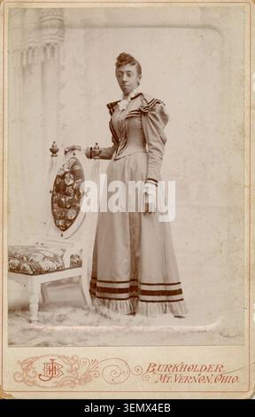 Vintage-Porträt einer Person, die neben einem Stuhl mit Blumenmuster steht, präsentiert in einem Schrankkartenformat von Burkholder in Mt. Vernon, Ohio. Stockfoto