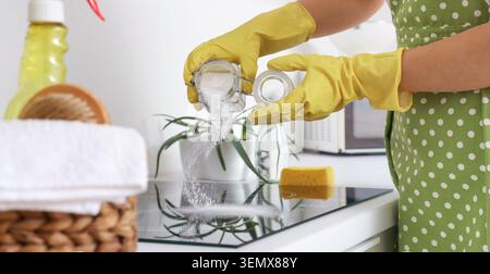 Frau in gelben Handschuhen, die weißes Pulver aus dem Glasbehälter auf ein schwarzes Induktionskochfeld für eine umweltfreundliche Reinigung gießt Stockfoto