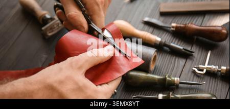 Handwerker schneiden rotes Leder mit einer Schere, handgefertigte Waren und maßgeschneiderte Accessoires auf einer Holzwerkbank, handwerkliches Können Stockfoto