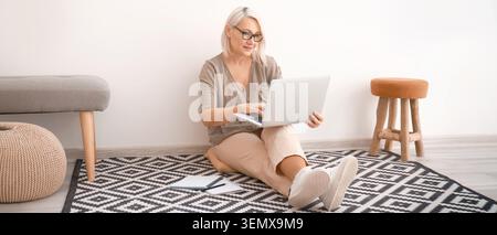Reife Frau, die von zu Hause aus mit einem Laptop arbeitet, Online-Lernen und Geschäftskommunikation nutzt, sitzt bequem auf einem gemusterten Teppich mit Notizbuch Stockfoto