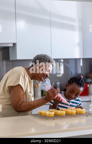 Die ältere Großmutter führt die Enkelin der Jugend, die rosa Glasur aus einer durchsichtigen Tüte auf Cupcakes in der Küche putzt Stockfoto