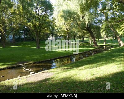 Ruhiger japanischer Garten im frühen Herbst mit einem ruhigen kleinen Fluss, der durch lebhaftes grünes Gras und üppiges grünes Laub fließt Stockfoto