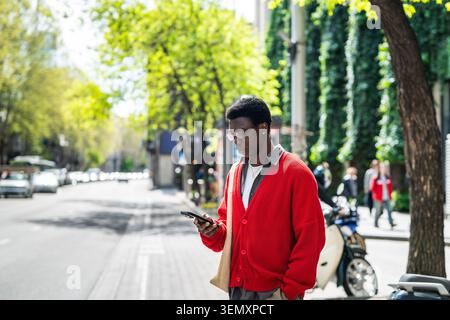 Entspannter afroamerikanischer Mann auf der Stadtstraße, der auf den Transport wartet und telefonisch den Fahrplan überprüft Stockfoto