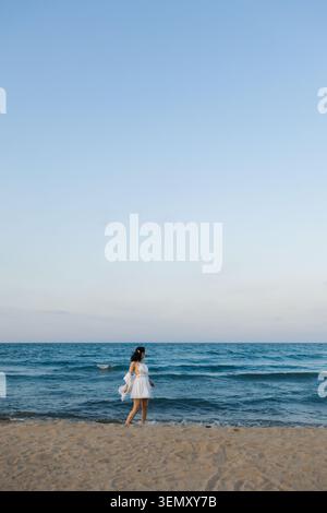 Eine Frau läuft am Sandstrand am Meer entlang, während die Sonne untergeht. Das Wasser reflektiert den Himmel, während die Frau ihre Zeit am Ufer genießt Stockfoto