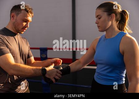 Männlicher Trainer passt die Handgelenkschlaufe an weibliche Athletin in blauem Oberteil im Boxring an, Smartwatch sichtbar Stockfoto