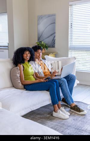 Ein Paar Afroamerikanerin, ein asiatischer Mann, der im Wohnzimmer auf dem Sofa sitzt, Laptop und Fernbedienung hält Stockfoto