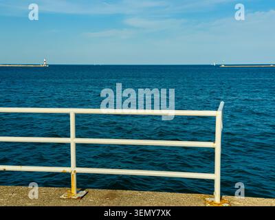 Die Szene zeigt einen Blick auf das ruhige Wasser von einem Pier. Ein weißes Geländer verläuft am Rand entlang. Im Hintergrund sind ein Leuchtturm und mehrere Boote zu sehen. Stockfoto