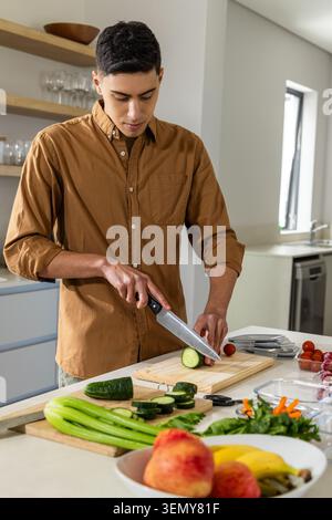 Mann in braunem Hemd, der auf der Kücheninsel steht und Gurke auf Schneidebrett mit Kochmesser schneidet Stockfoto