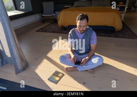 Reifer asiatischer Mann, der auf dem Schlafzimmerboden neben der Glastür sitzt und ein Smartphone hält, Taschenbuch in der Nähe Stockfoto