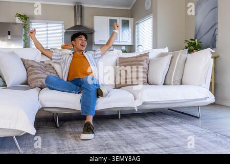 Asiatischer Mann in seinen Zwanzigern, der auf weißem Sofa sitzt und Arme, Laptop und Orangen in der Nähe hochhebt Stockfoto