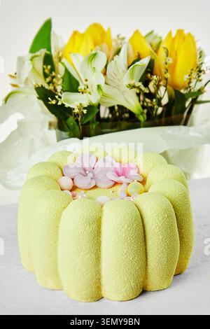 Gelber Mousse-Kuchen mit Zuckerblumen auf weißem Tisch mit verschwommenem Frühlingsstrauß im Hintergrund. Elegantes festliches Dessert mit Kopierraum. Stockfoto