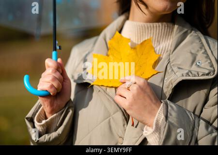 Konzeptionelle Herbstszene, die einen Teil des Körpers der Frau zeigt, eine Hand den Schirmgriff hält, die andere Hand das gelbe Ahornblatt in Brusthöhe hält, Highlight Stockfoto