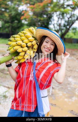 Bananenfrau Hut Lächeln tropische Frucht Farm Ernte im Freien rotes Hemd Junge Frau hält Bananenbunch auf der Schulter, trägt Strohhut mit glücklichem Lächeln Stockfoto