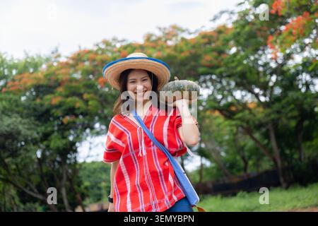 Frau Hut Kürbis rotes Hemd Garten Lächeln Natur Baum Blume im Freien fröhliche junge asiatische Frau trägt Strohhut und rot gestreiftes Hemd, das grüne p hält Stockfoto