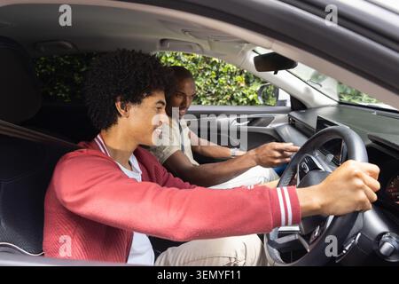 Männlicher Lerner in roter Jacke, der das Rad dreht, während der Kursleiter auf den Armaturenbrett im Auto zeigt, Sicherheitsgurte angelegt Stockfoto