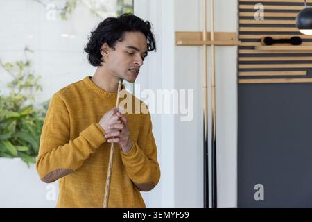 Asiatischer Mann, der Billardkurve hält, Kinn in der Nähe von Tip im Spielzimmer mit Senfpullover Stockfoto