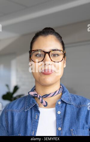 Frau mit Schildkrötenbrille gemustertem Halstuch und Jeansshirt lächelt beim Posen im Büro Stockfoto
