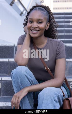 Afrikanerin sitzt auf städtischen Stufen mit lächelndem Handlauf, trägt T-Shirt, Jeans, braune Umhängetasche Stockfoto