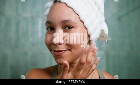 Frau, Die Im Badezimmer Mit Handtuchumwickelten Haaren Befeuchtet, Lächelnde Hautpflege-Routine Stockfoto