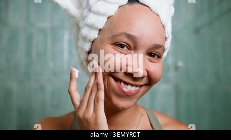 Smiling Woman Wendet Gesichtscreme Mit Handtuchumwickelten Haaren Während Der Morgendlichen Hautpflege-Routine An Stockfoto