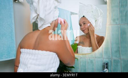 Schwarze Frau, die Hautpflege-Routine im Badezimmerspiegel mit Handtuch Turban und Natural Glow macht Stockfoto