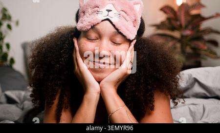 Entspannte Frau lächelnd im Bett trägt rosa Katze Schlafmaske in gemütlichem Schlafzimmer Morgen Selbstversorgung Stockfoto