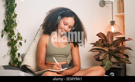Schwarze Frau, die zu Hause mit Kopfhörern in gemütlichem Schlafzimmer mit Pflanzen und Lampe arbeitet Stockfoto