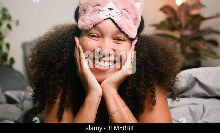Lächelnde Frau Mit Rosa Katze Schlafmaske Entspannt In Gemütlichem Schlafzimmer Porträt Mit Lockigem Haar Stockfoto