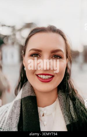 Frauenporträt Lächeln mit einem jungen Erwachsenen mit rotem Lippenstift, der draußen in einer urbanen Umgebung einen karierten Schal trägt Stockfoto