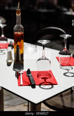 Eine Nahaufnahme eines weißen Restauranttisches mit umgedrehten Weingläsern, roten Servietten, Besteck und einer Flasche Chili-Öl. Stockfoto