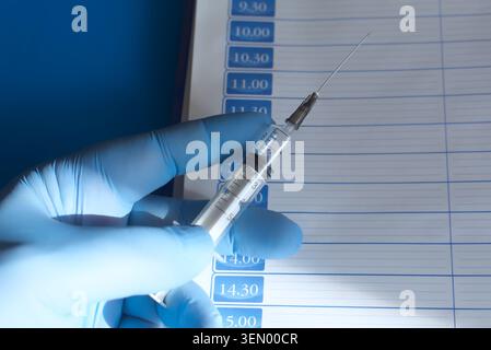Hand in einem blauen medizinischen Handschuh, der eine medizinische Spritze im Sonnenlicht über eine Seite eines Tagesplaners hält, Nahaufnahme. Stockfoto