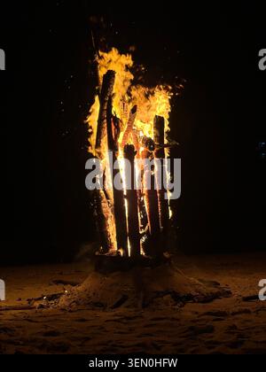 Ein Strandfeuer lodert gegen die schwarze Nacht, seine aufrecht stehenden Holzblöcke glühen wie eine feurige Skulptur im Sand Stockfoto
