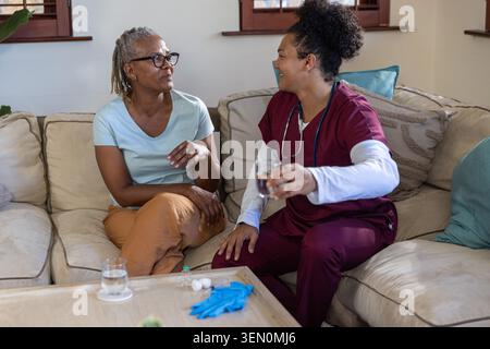 Seniorin und mittelerwachsene Pflegekraft, die auf der Couch sitzen und Wasser anbieten, mit Stethoskop Stockfoto