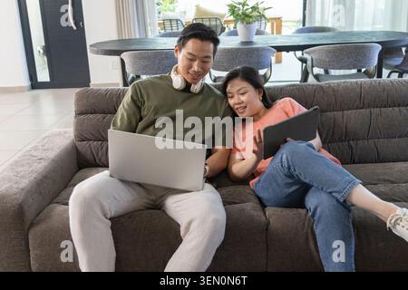 Asiatisches Paar, das im modernen Wohnzimmer auf einem grauen Sofa sitzt, mit silbernem Laptop und dunklem Tablet Stockfoto
