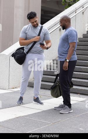 Verschiedene männliche Freunde in Sportbekleidung teilen ihr Smartphone, während sie auf plaza Steps Reisetaschen halten Stockfoto