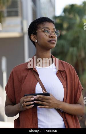 Afroamerikanerin, die auf der Stadtstraße steht und ein Smartphone hält, Rosthemd und Brille trägt Stockfoto