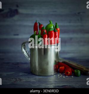 Rote und grüne Chilischoten in einem Edelstahlbecher auf rustikalem Holztisch mit geschnittenen Paprika, Samen und Vintage-Messer. Stockfoto