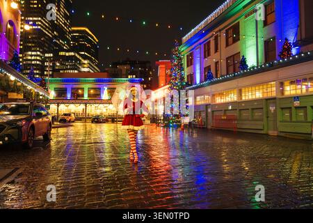 Eine Person in festlichem Outfit steht auf einer nassen, beleuchteten Straße, die mit bunten Lichtern dekoriert ist. Seattle, USA Stockfoto