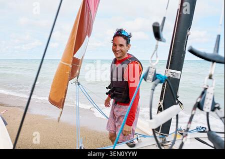 Lächelnder Mann in Segelausrüstung, der an einem sonnigen Tag in der Nähe eines Segelbootes steht. Stockfoto