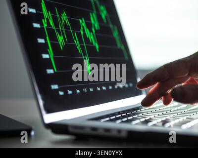 Hand zeigt auf den Laptop-Bildschirm mit grünem Finanzdiagramm in einem schwach beleuchteten Raum. Stockfoto