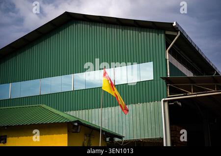 Die Staatsflagge von Selangor winkt vor einem großen Industrielager aus grünem Wellblech Stockfoto