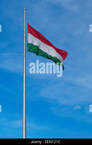 Flacher Blick auf die Nationalflagge Ungarns, die auf einem hohen Metallfahnenmast vor einem blauen Himmel mit hellen Wolken winkt. Stockfoto